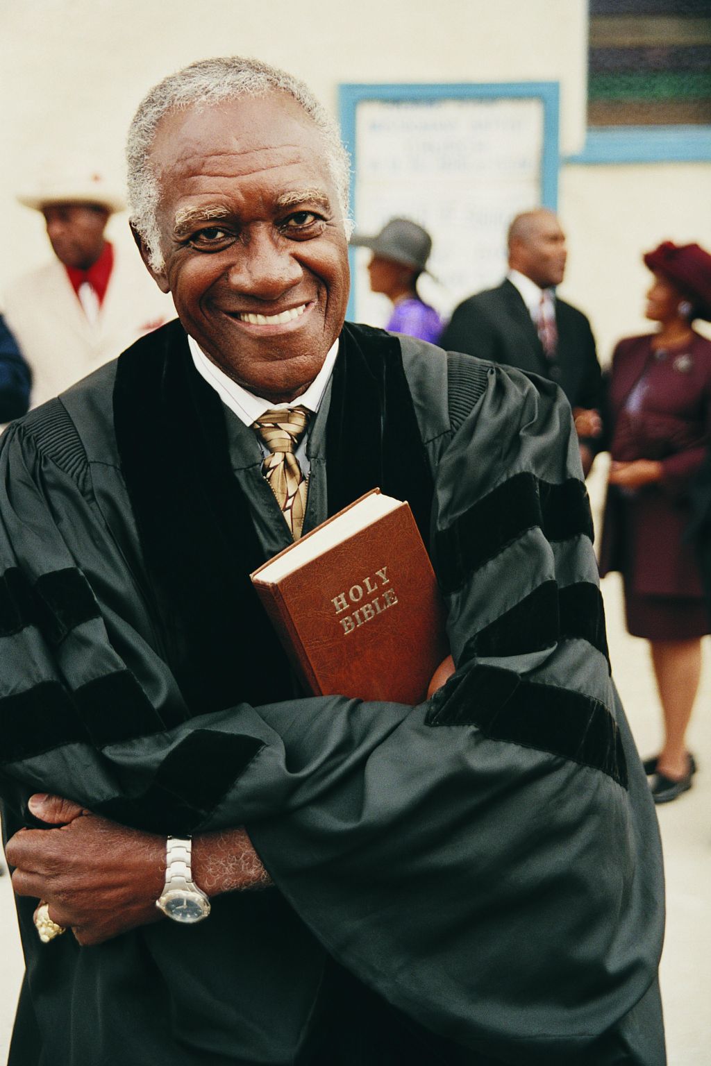 Portrait of a Smiling Priest Holding a Bible in His Crossed Arms With People in the Background