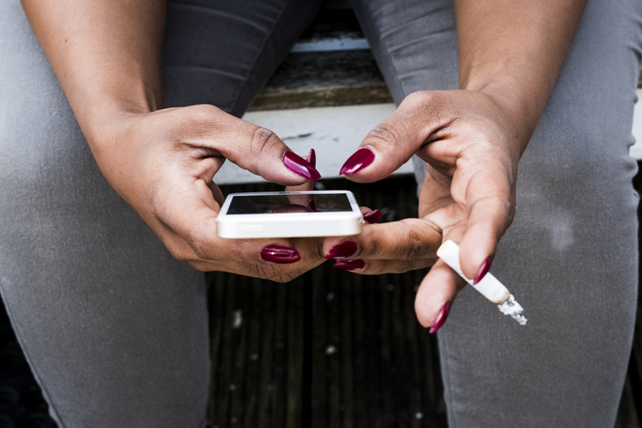 A woman, hand holding a cigarette and texting on her smart phone.