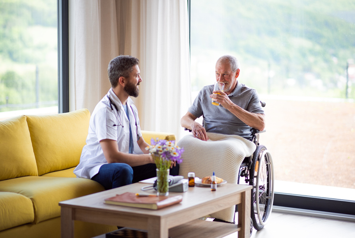 A male healthcare worker talking to senior man in wheelchair indoors at home.