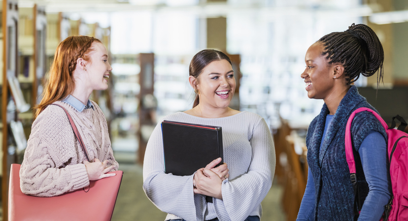 Three teenage girls standing in library conversing