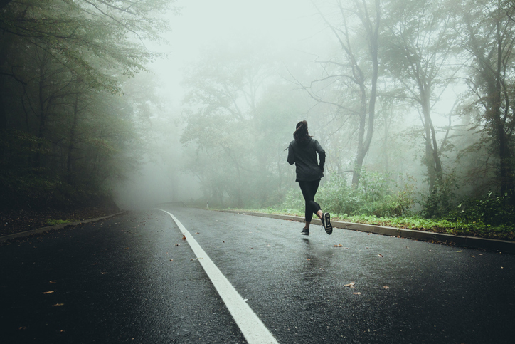 Rear view of female athlete running on the road in misty forest.