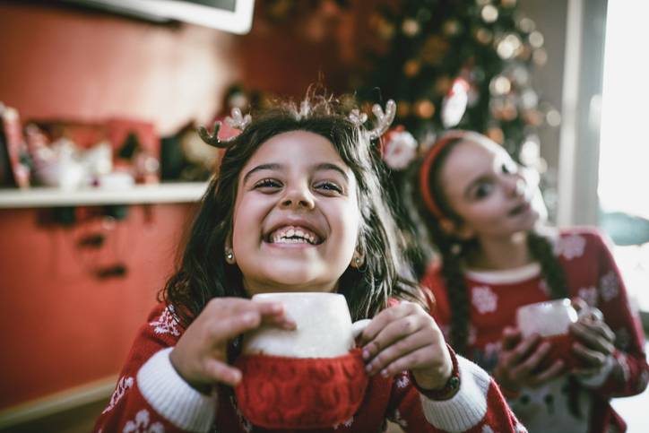 Two Little Sisters Playing Around Christmas Tree and Drinking Hot Beverage