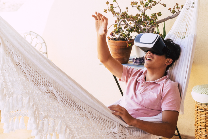 happy teenager having fun with his new VR glasses in the terrace of his house on an hammock - adult tryng to touch something he see in the glasses of the future