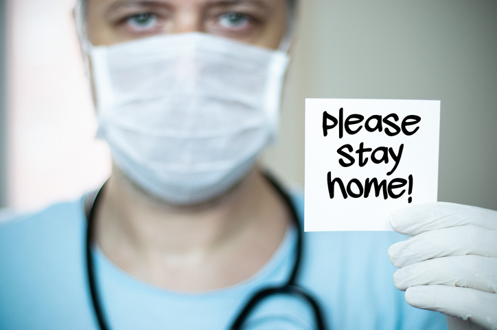 The middle-aged, green-eyed doctor with a mask on his face and holds a paper that says "please stay home"